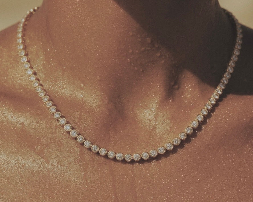 A close-up of a delicate diamond tennis necklace worn on a model's neck, with warm golden lighting and a soft-focus background.