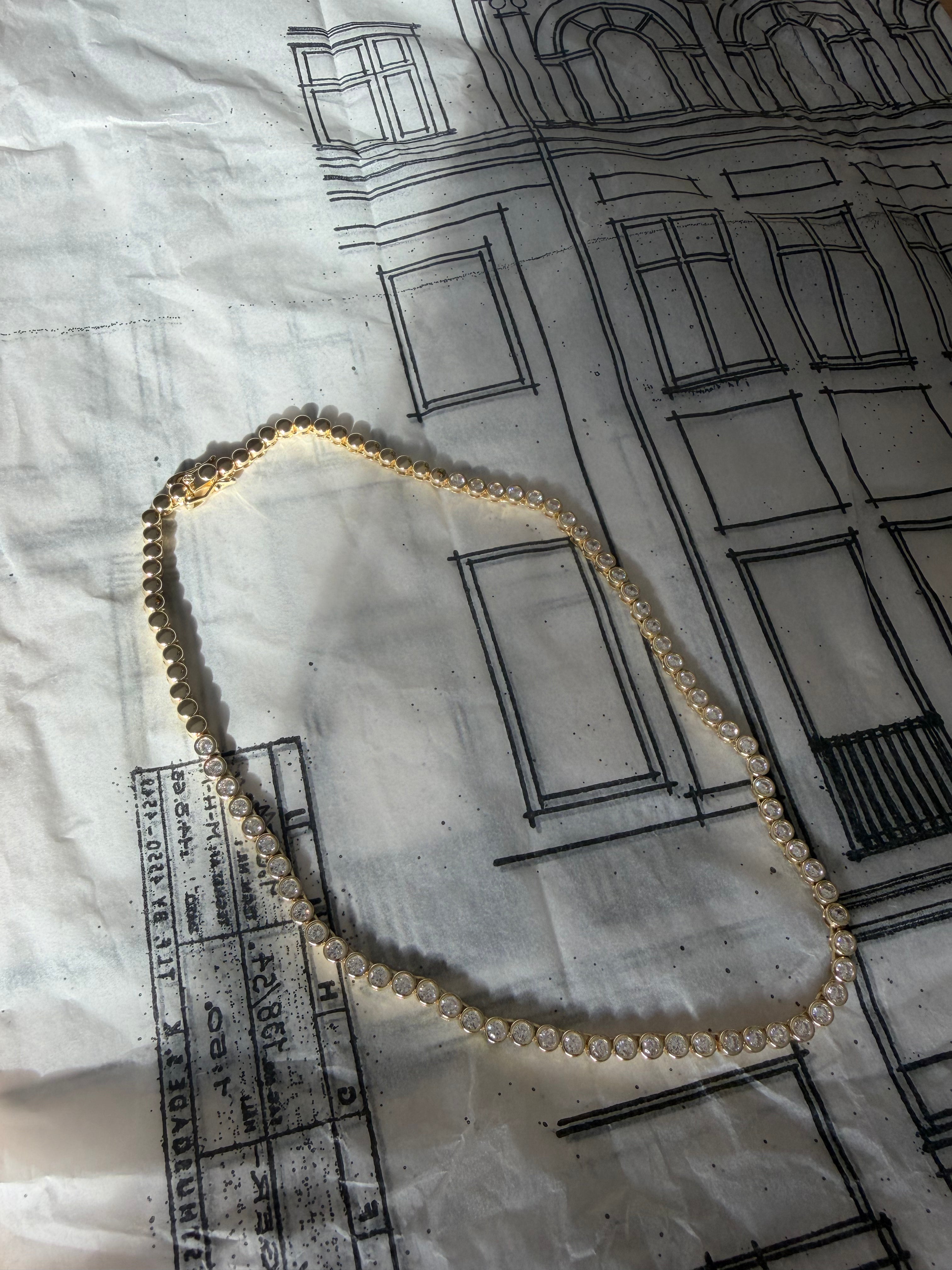 A gold chain necklace with small clear stones, arranged in a loop on a white paper background printed with black architectural building sketches.