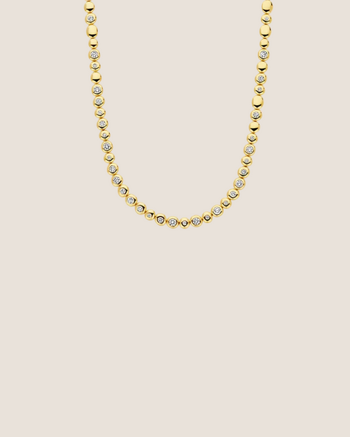 A gold tennis necklace featuring small round bezel-set clear stones, shown against a plain white background.