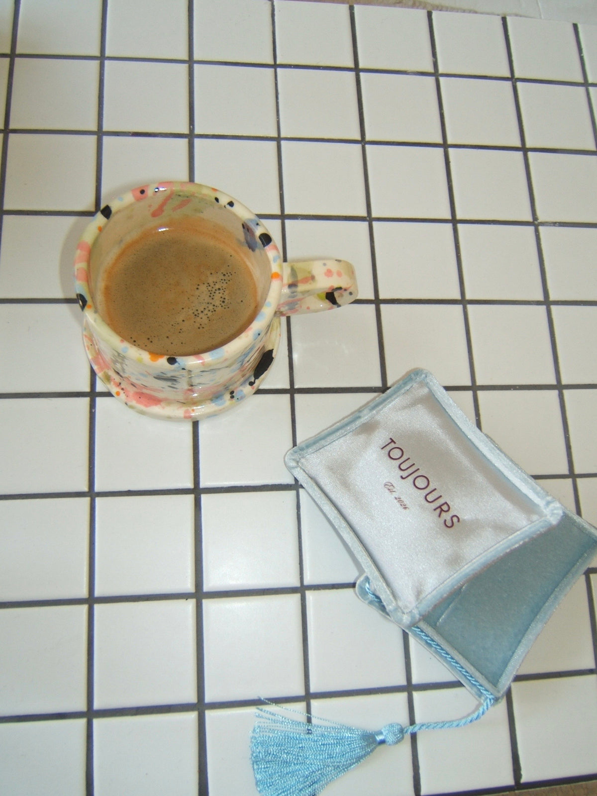A small light blue velvet pouch with a tassel and the word 'TOIJOURS' printed on it, placed on a white tiled surface next to a patterned mug containing coffee.